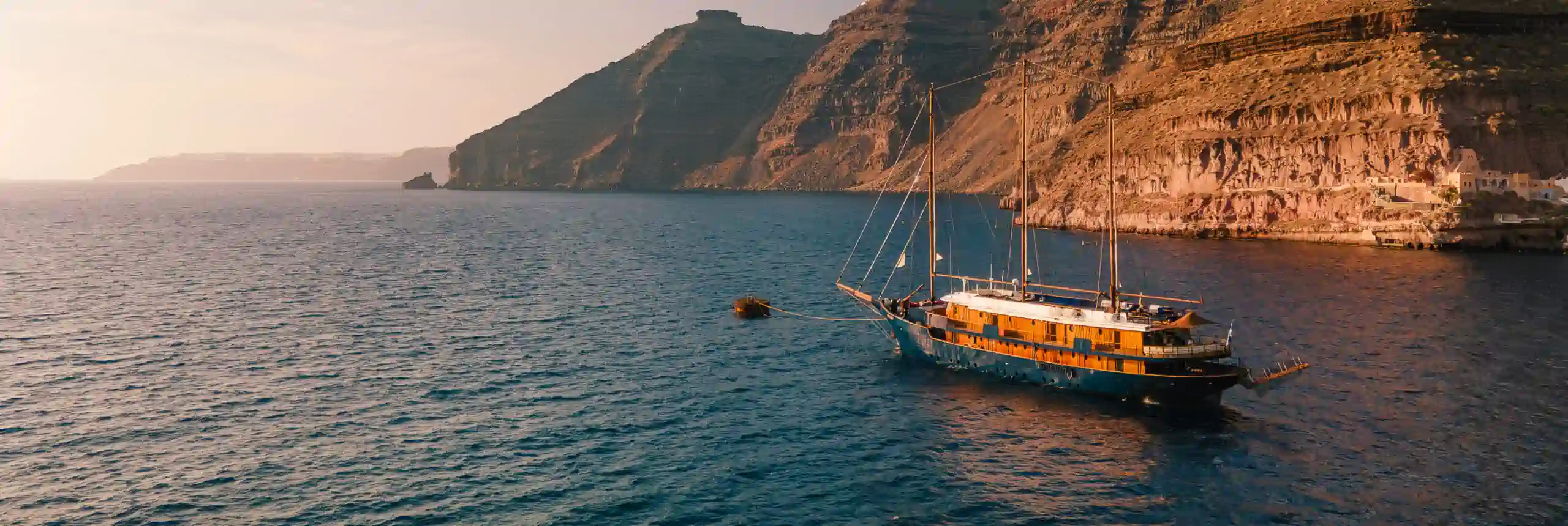 Wooden motor sailer at anchor beneath the cliffs of Santorini at golden hour