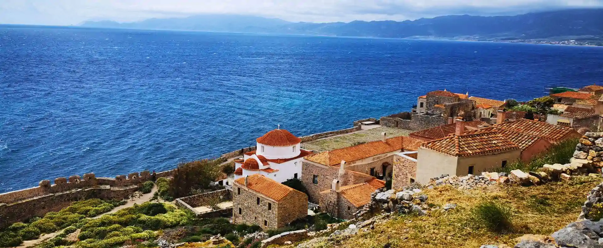 cyclades-cruise-itinerary-monemvasia-sea-panorama.webp Panoramic Aegean view on a Cyclades cruise itinerary featuring red-roofed chapel and coastal Monemvasia set above vivid blue sea.