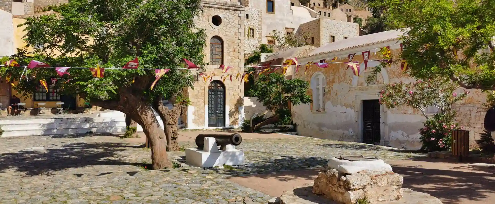 cyclades-cruise-itinerary-monemvasia-village-square.webp Traditional square visited on a Cyclades cruise itinerary, with bunting, olive tree and historic church walls in Monemvasia.