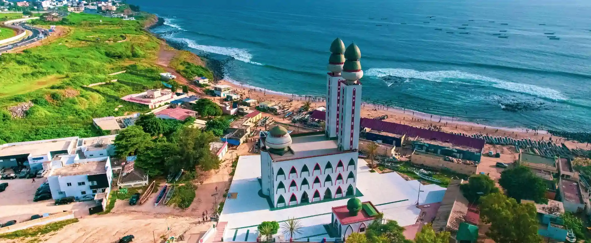west africa cruises senegal coastline mosque Aerial view of a colourful mosque on the Senegal coastline, with waves, fishing boats, and lush green hills.