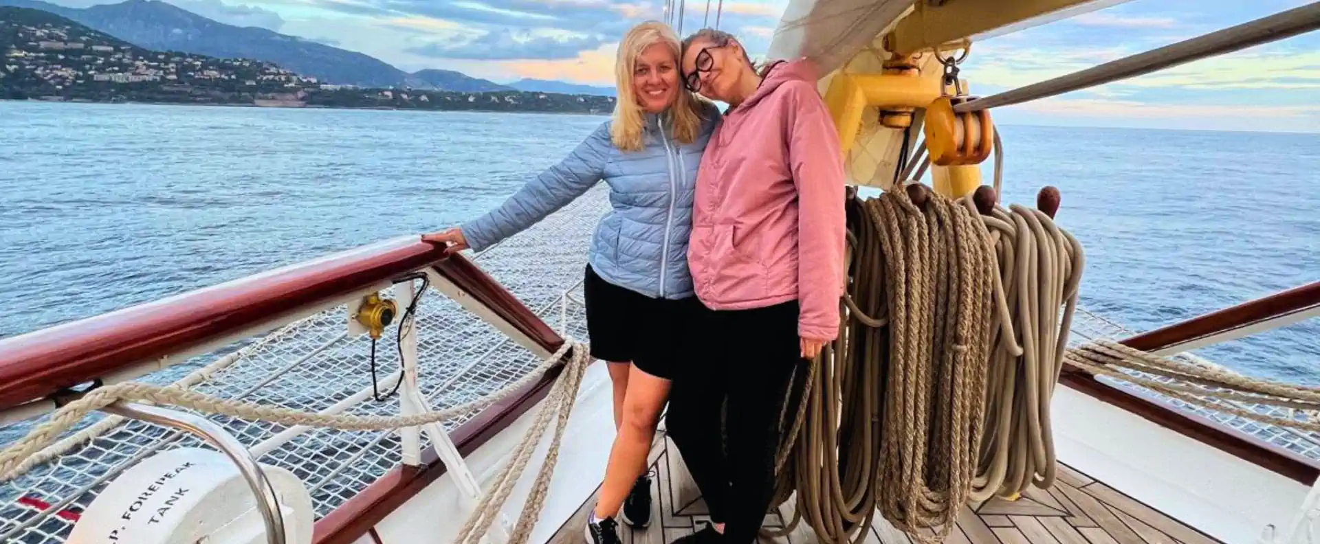 Guests on Star Flyers bowsprit netting at sunset with coastal hills in the background.